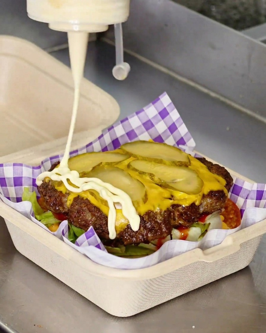 White mayonnaise being squeezed from a bottle in a zigzag pattern over a cheeseburger patty.