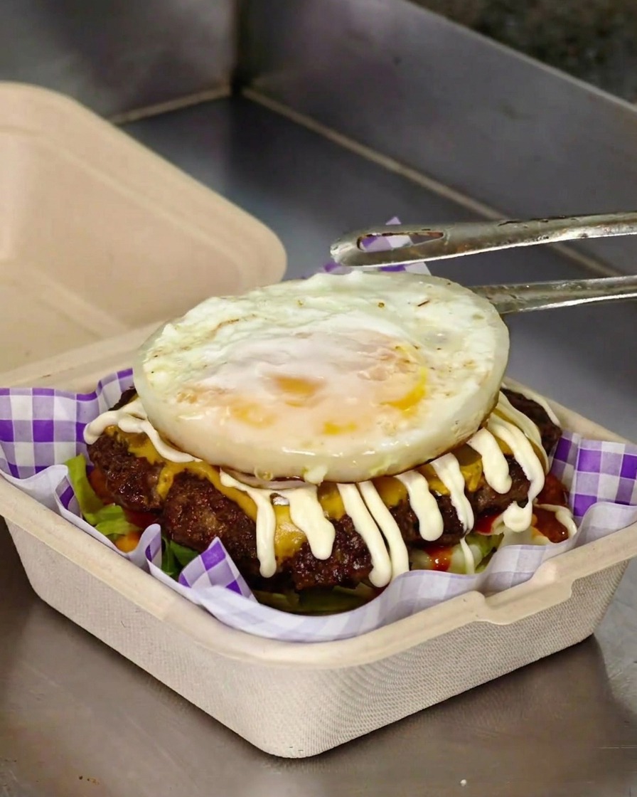 Metal tongs placing a freshly fried egg on top of a burger loaded with smashed meat patties, melted cheese, and a zig-zag drizzle of mayonnaise.