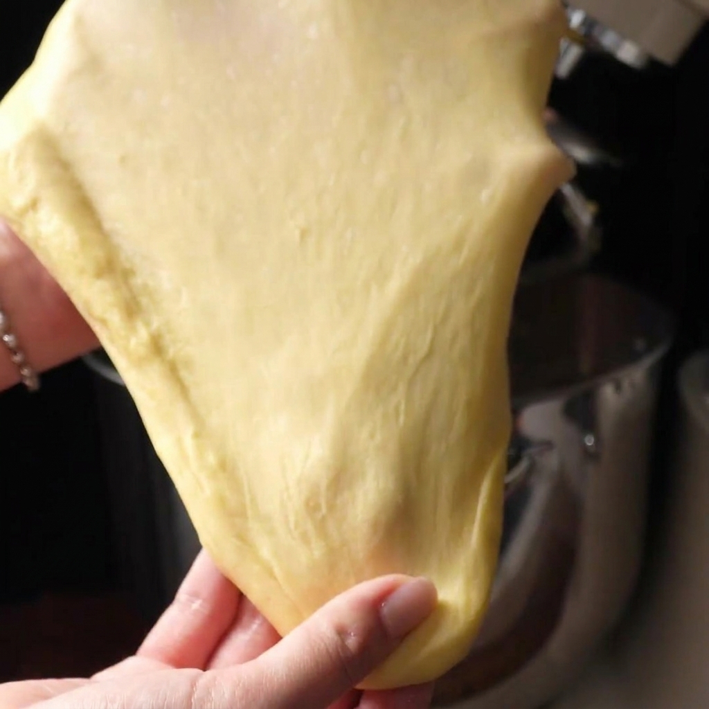 Two hands gently stretching a piece of smooth dough into a very thin, translucent film known as a windowpane.