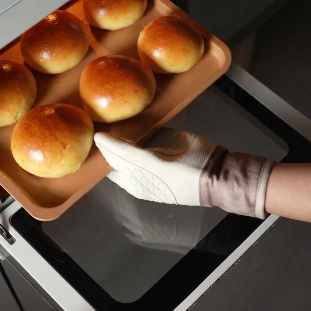 A hand wearing a white oven mitt pulling a copper baking tray filled with perfectly golden brioche buns out of a hot oven.