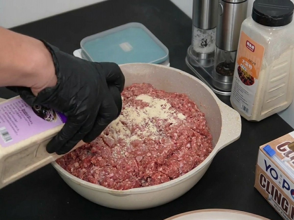 A gloved hand sprinkling onion powder from a container over a pan of raw ground beef.