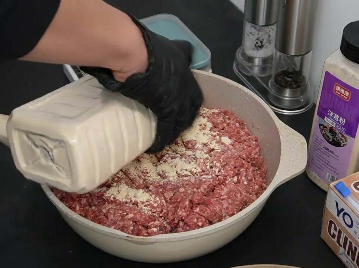 A person wearing black gloves pouring white garlic powder from a large container over a pan of raw ground beef.