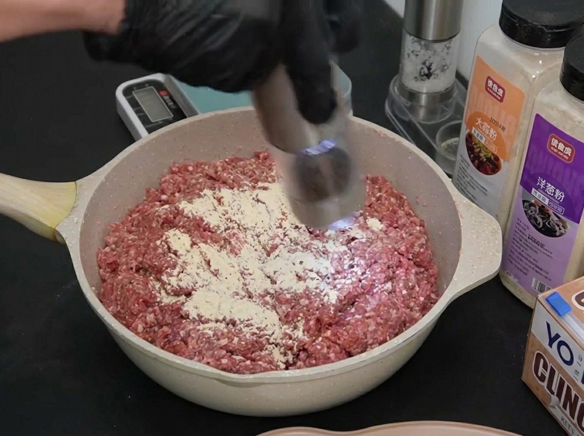 A gloved hand using an electric grinder to disperse black pepper over seasoned raw ground beef.