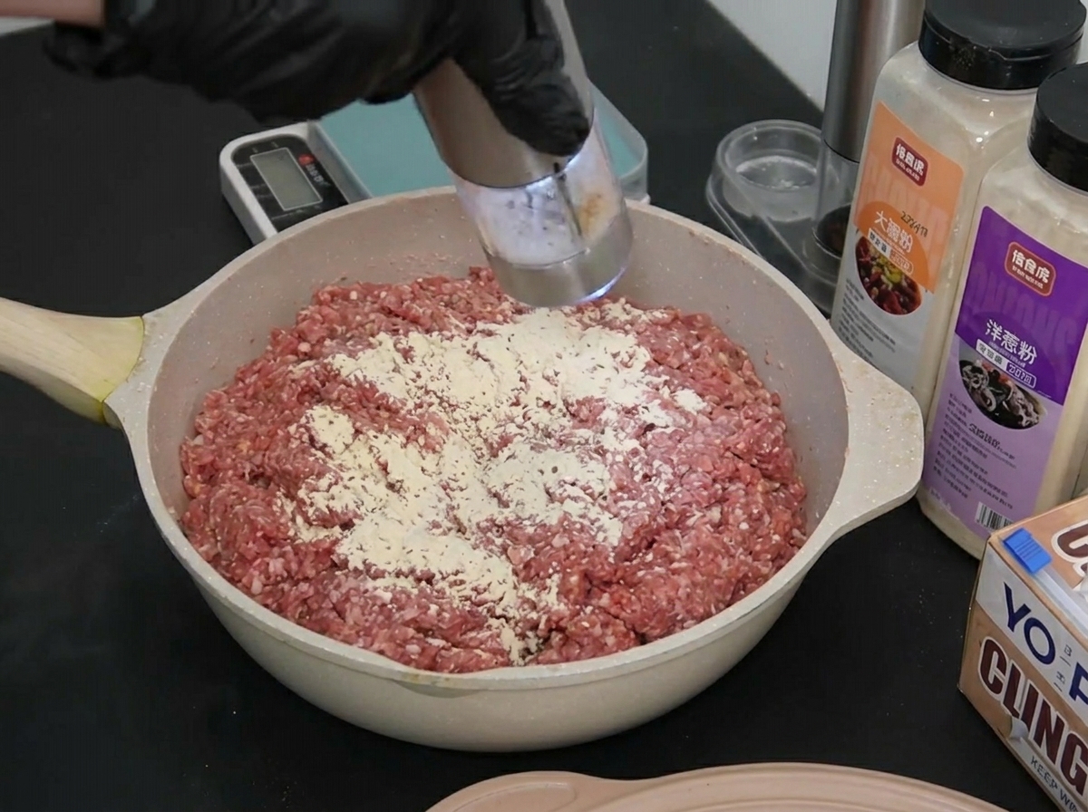 A gloved hand grinding a small amount of white sea salt onto the surface of seasoned raw ground beef.
