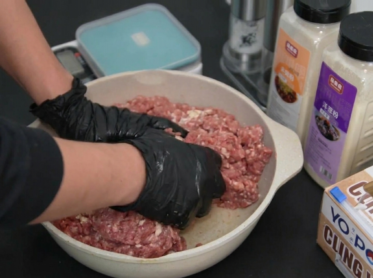 Two gloved hands pressing into and mixing raw ground beef inside a shallow pan.