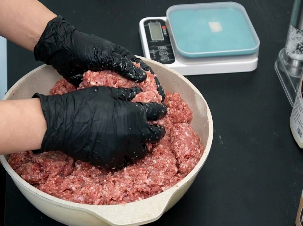 Two gloved hands lifting a portion of mixed and seasoned raw ground beef from a pan.