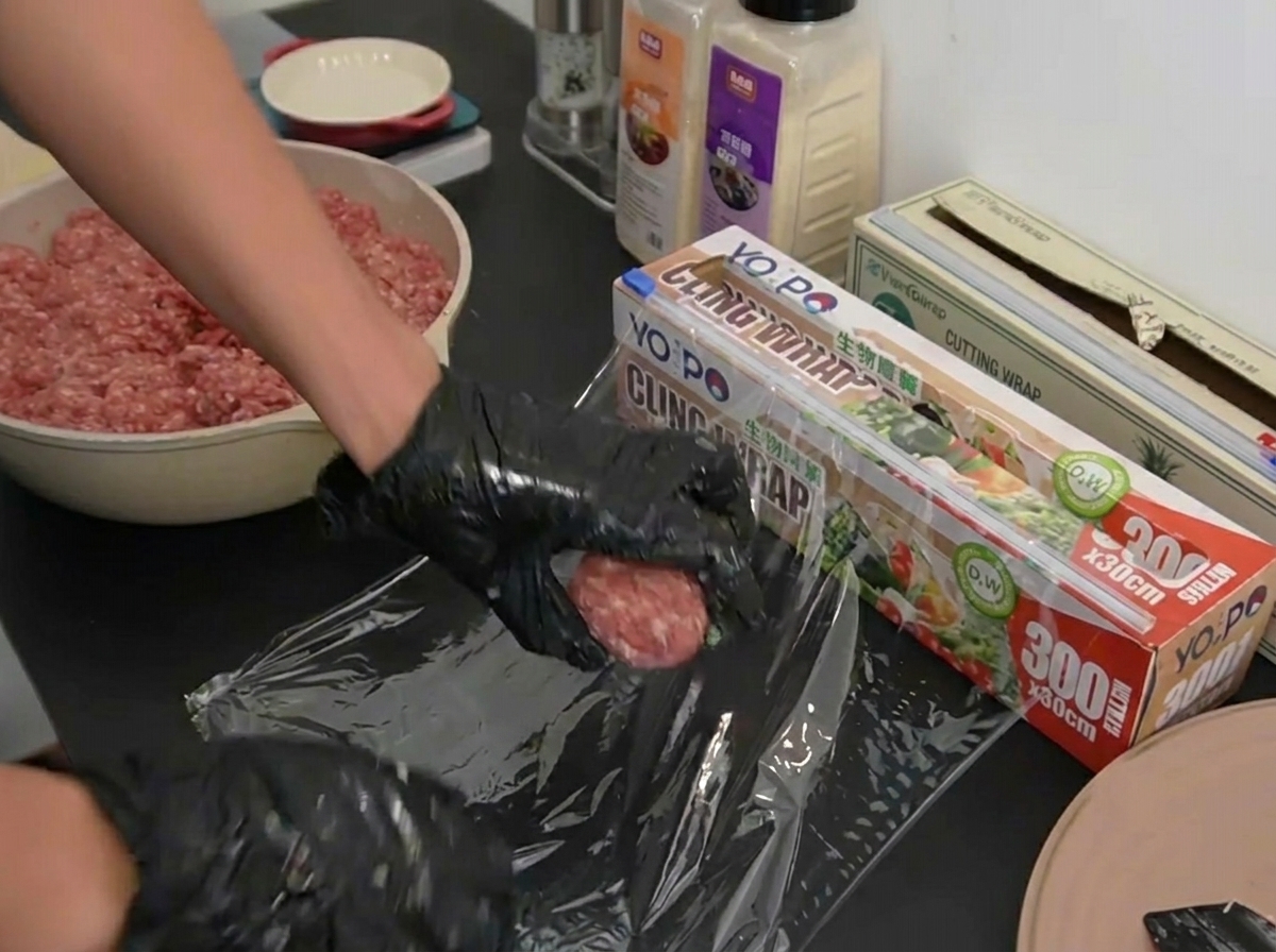 Gloved hands gently placing a perfectly portioned, raw beef ball onto a clear sheet of laid-out plastic wrap.