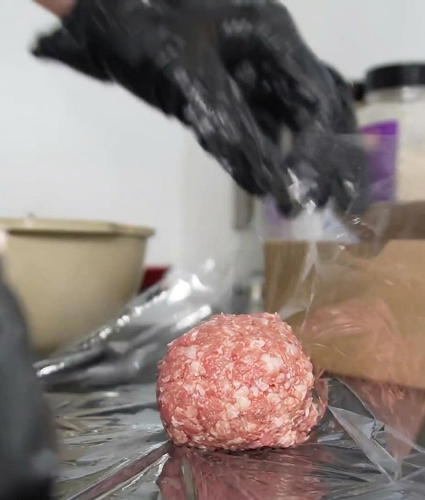 Gloved hands tightly twisting clear plastic wrap around a raw beef patty to create a sealed package.