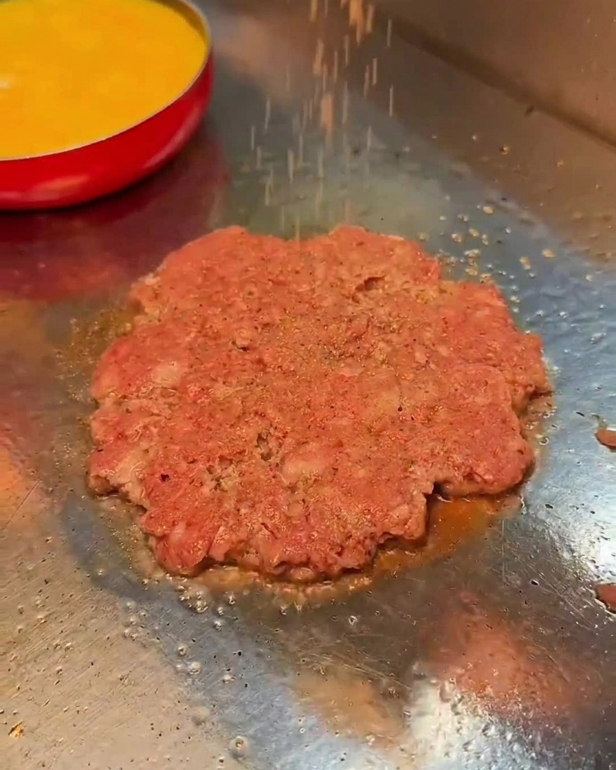 Salt and black pepper being sprinkled over a cooking beef patty on a hot flat-top grill.