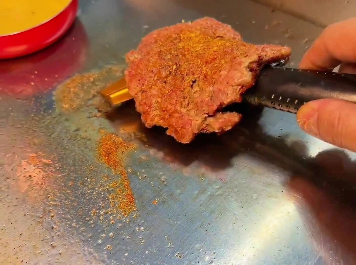 A smashed beef patty turned over on a grill, revealing a deeply browned and seasoned crust.