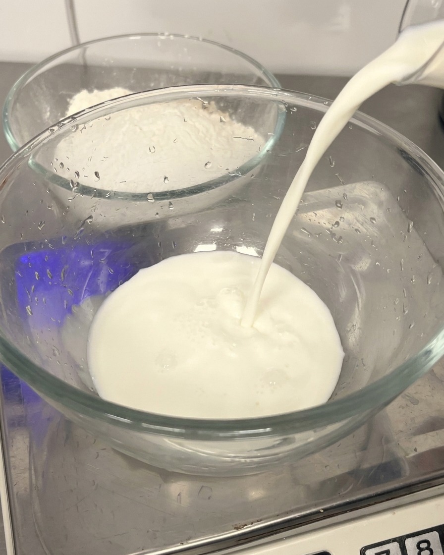 Fresh milk being poured into a clear glass bowl, with a bowl of flour visible in the background.