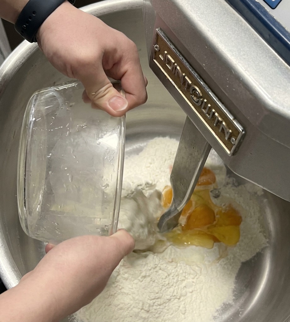 A stand mixer dough hook incorporating eggs and liquid into a pile of flour inside a large stainless steel bowl.