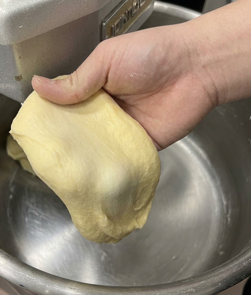 A hand stretching a piece of pale yellow brioche dough to demonstrate the thin, strong windowpane membrane.