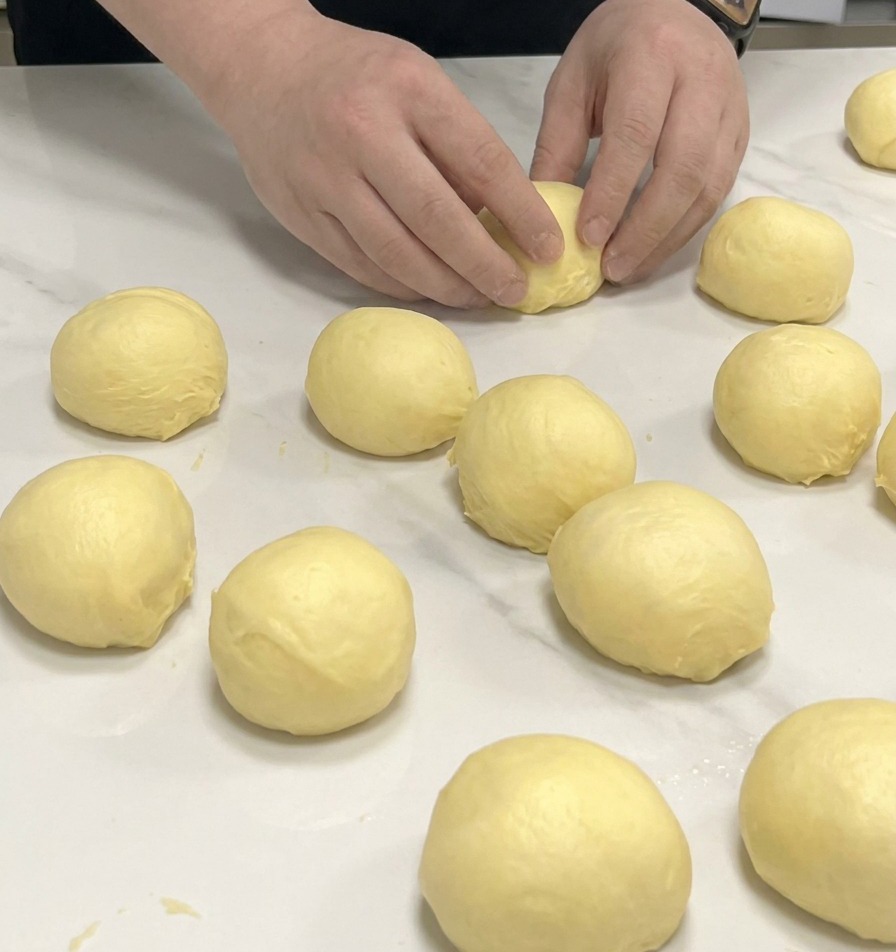 Hands rounding small portions of yellow brioche dough into balls on a white marble countertop.