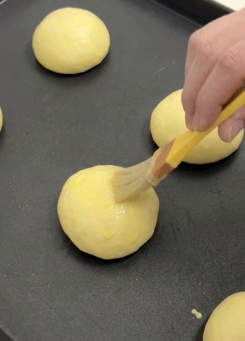 A yellow pastry brush applying a thin layer of egg wash to the top of a proofed brioche bun on a baking tray.