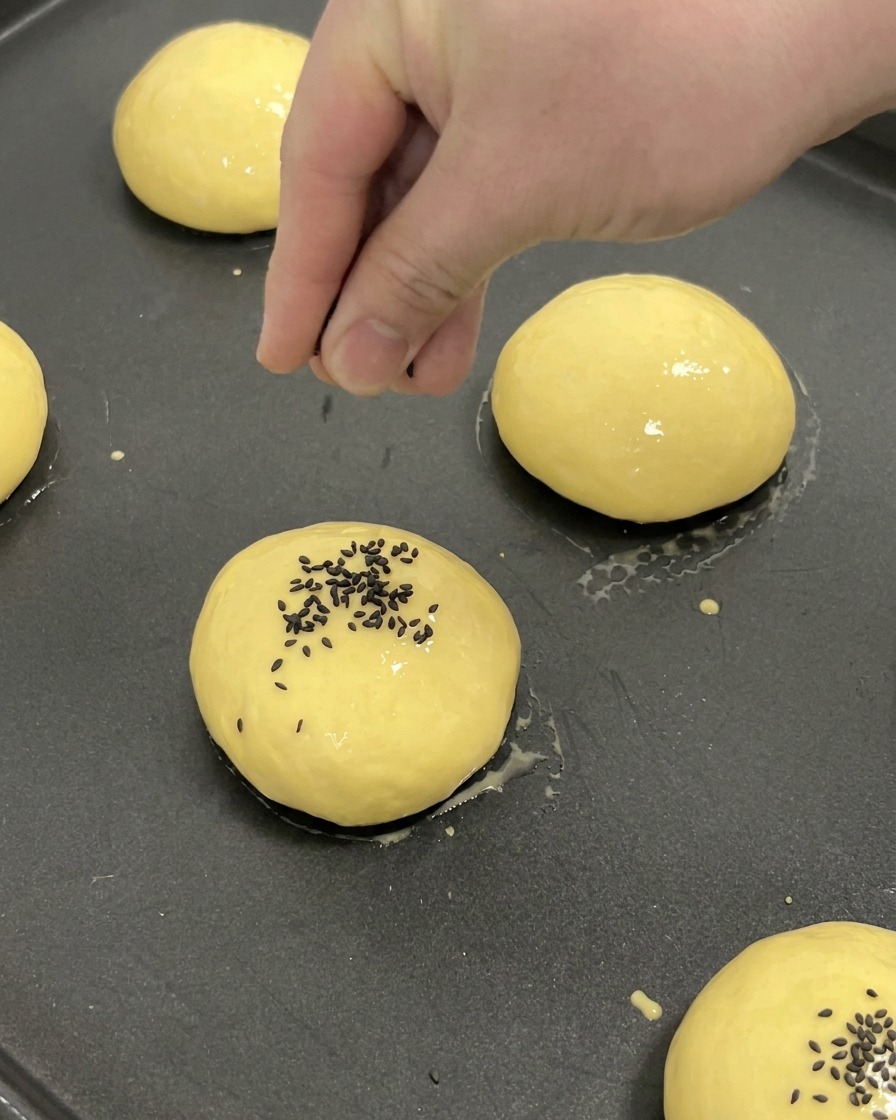 A hand sprinkling black sesame seeds onto the center of a proofed, egg-washed brioche bun on a baking tray.
