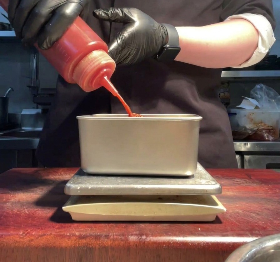 Gloved hands squeezing bright red sriracha sauce from a bottle into a metal prep container.