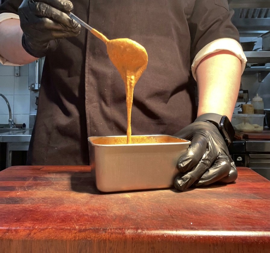 A gloved hand lifting a spoonful of smooth, vibrant orange burger sauce from a metal container.
