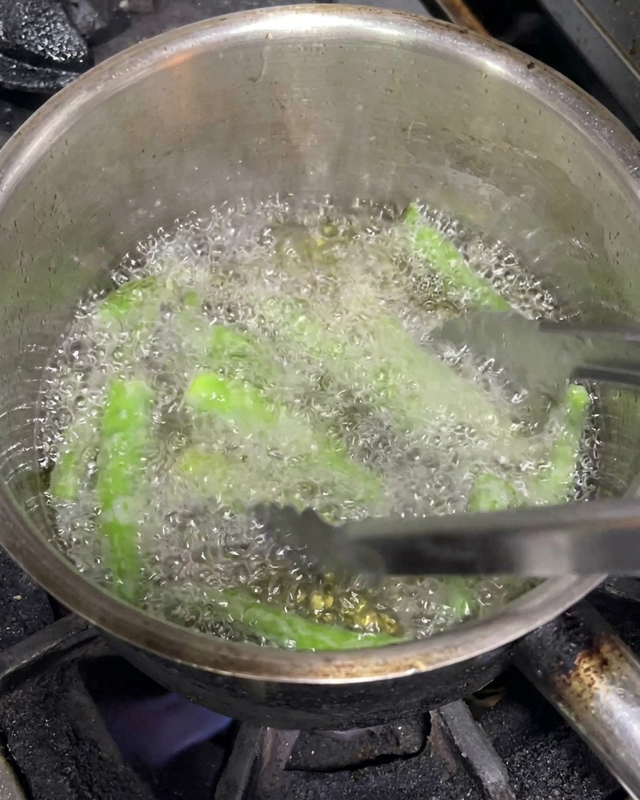 Metal tongs maneuvering whole green chilies as they deep-fry in a pot of bubbling hot oil.