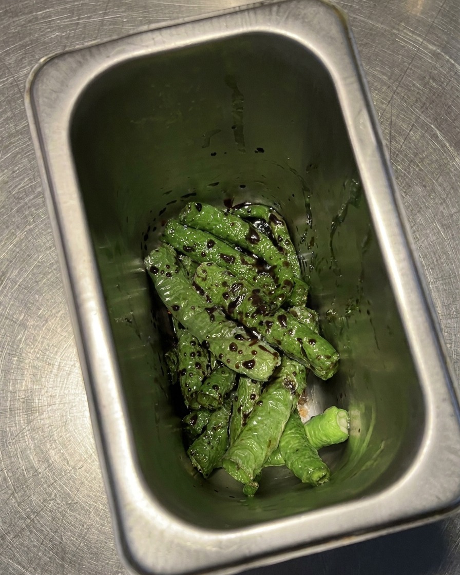 Fried, blistered green chilies sitting in a metal container, heavily coated in a dark mixture of vinegar and soy sauce.