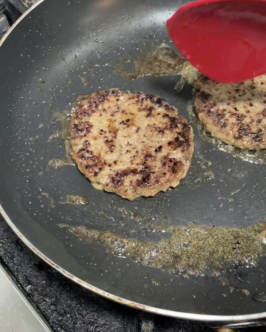 Two thick beef patties sizzling in a hot flat pan, developing a nicely browned crust with a red spatula resting on one of them.