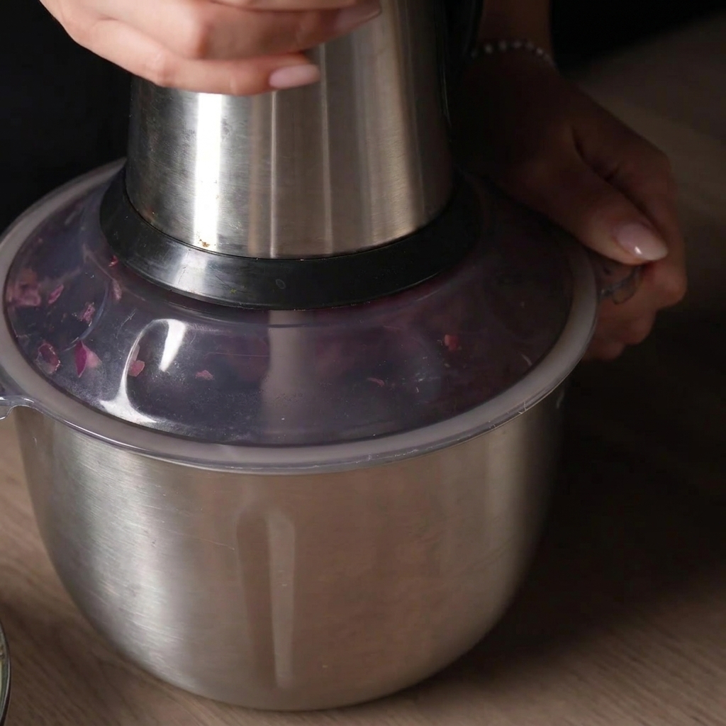A hand pressing down on the lid of a metal food processor to pulse the raw meat mixture inside.