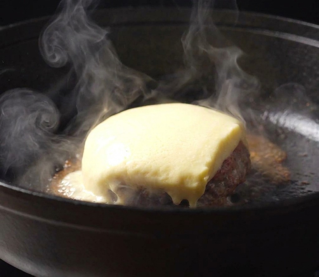 A thick slice of pale yellow cheese melting over a beef patty in a hot, steaming cast-iron skillet.