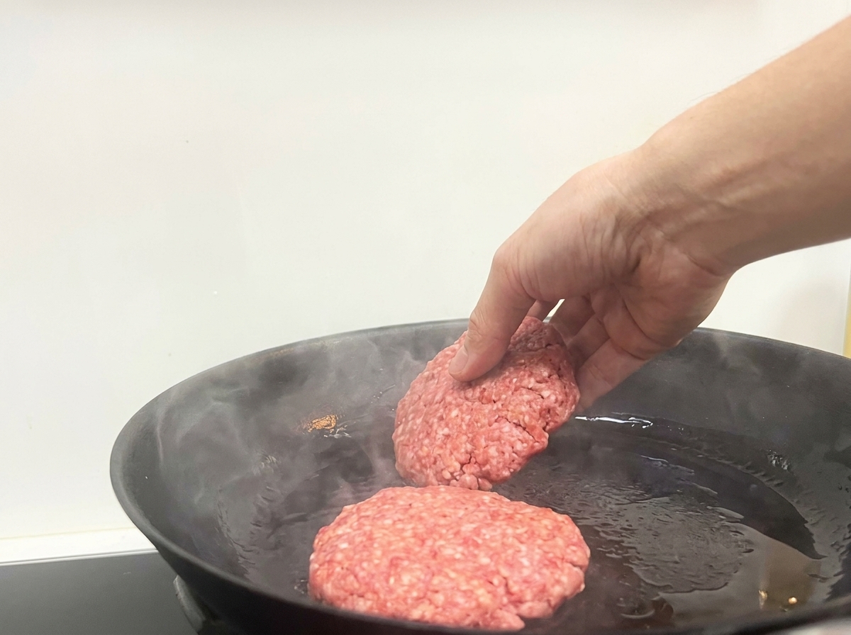 A hand placing two raw ground beef patties into a hot, slightly smoking black frying pan.