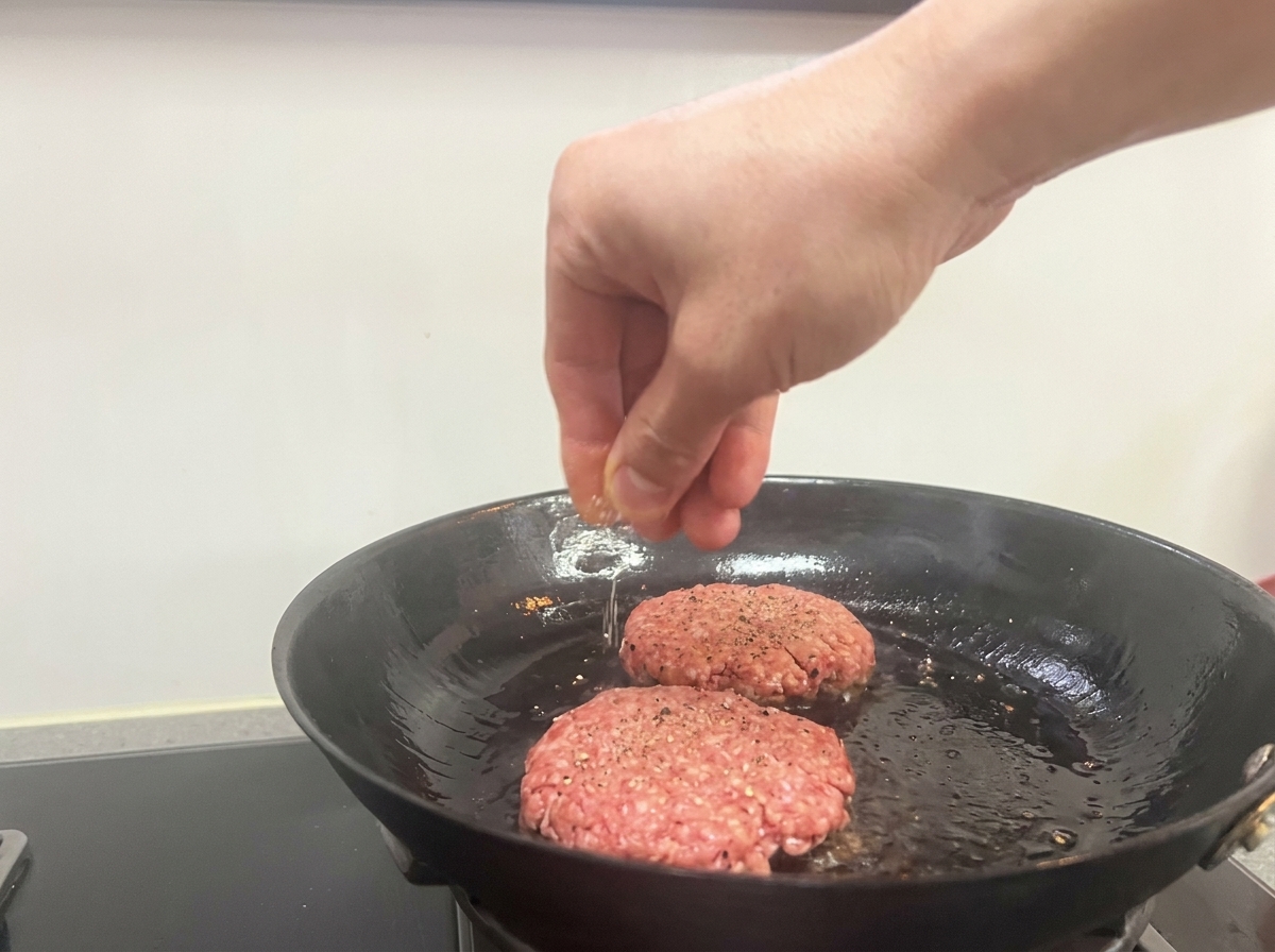 A hand sprinkling salt and black pepper over two searing beef patties in a frying pan.