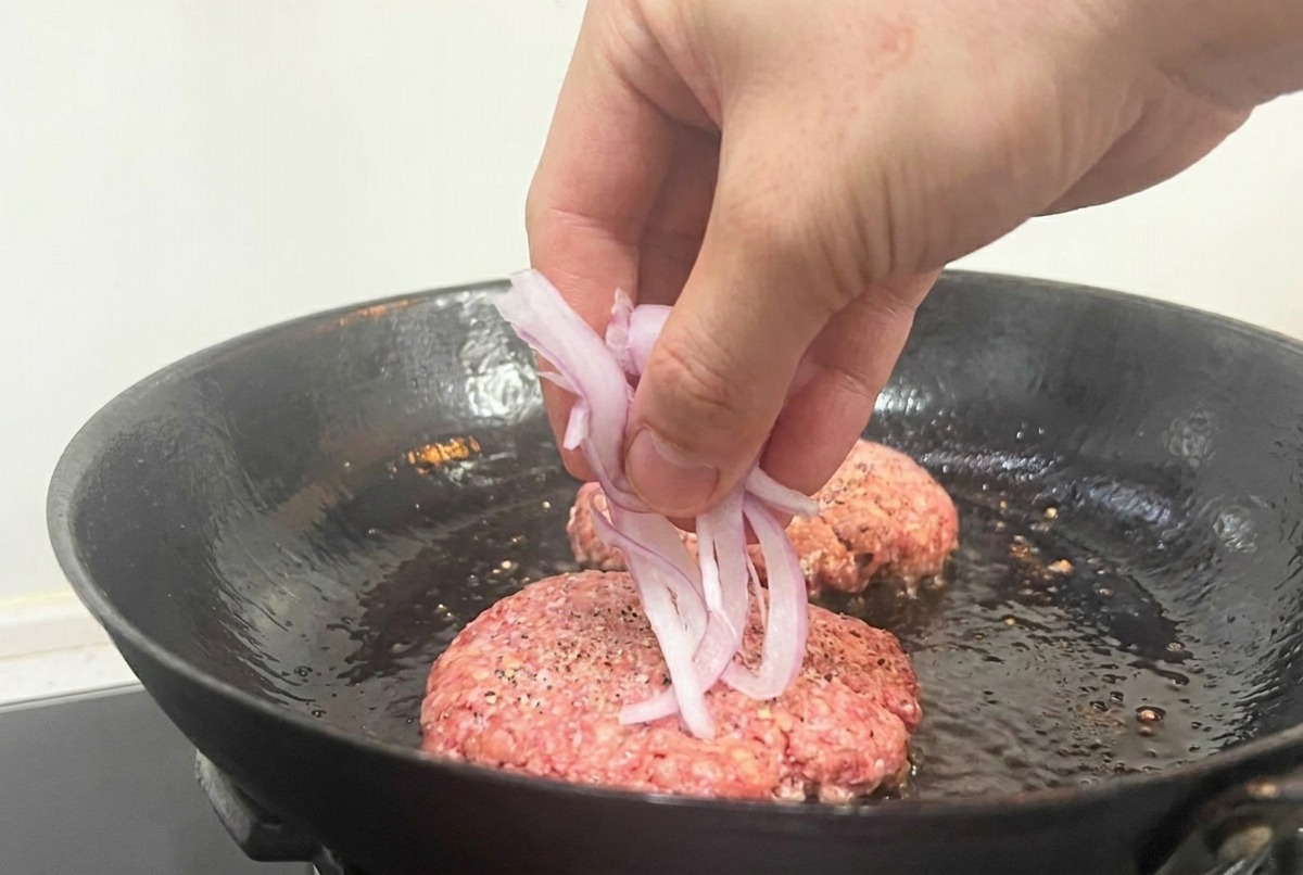 A hand dropping sliced red onions onto raw beef patties cooking in a black skillet.