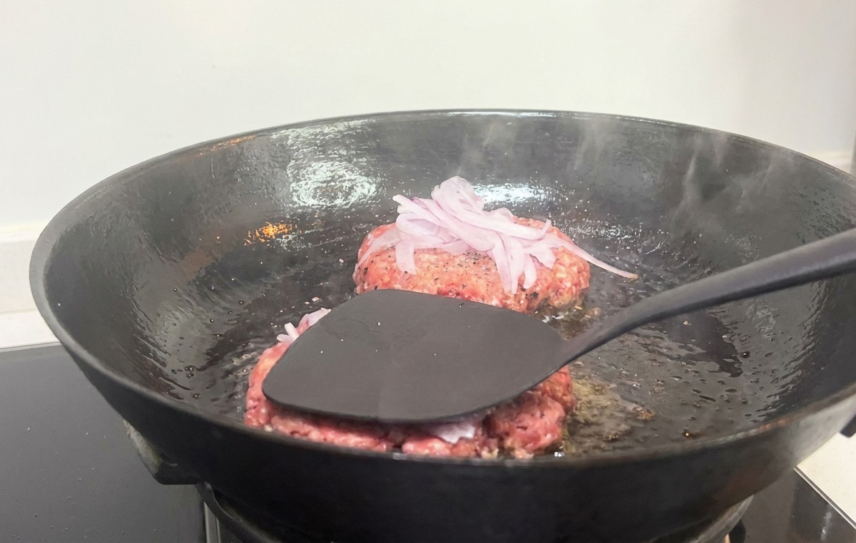 A black spatula firmly pressing down on a burger patty with sliced red onions in a hot cast iron pan.