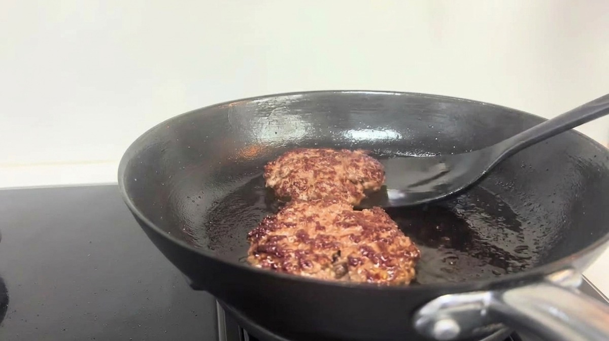 A black spatula turning over browned beef patties in a frying pan.