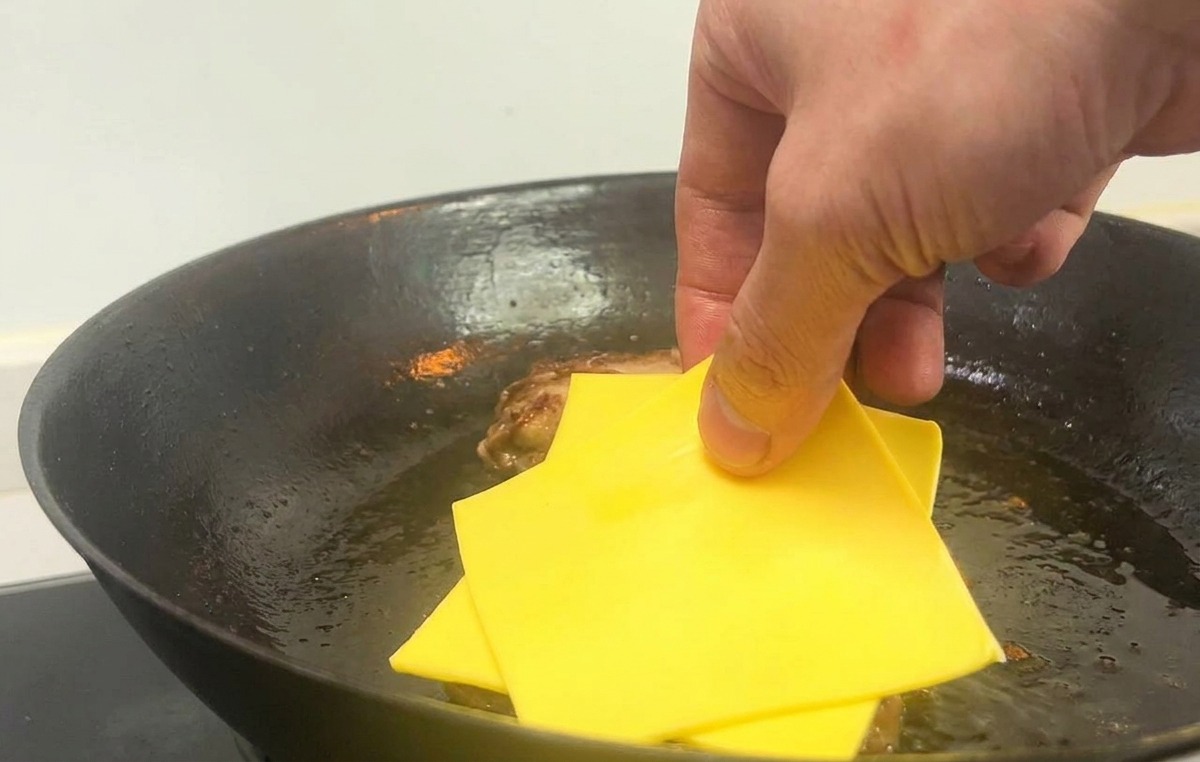A hand placing two square slices of yellow cheese onto cooked burger patties in a hot skillet.