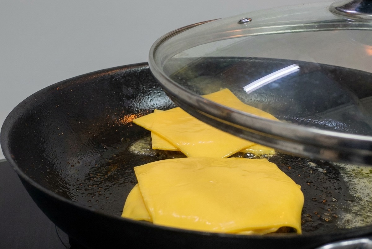 A glass lid being placed over a pan containing burger patties topped with melting yellow cheese.