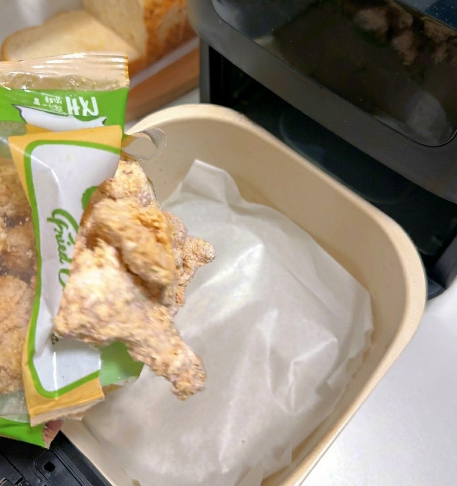 Frozen breaded chicken pieces being emptied from a bag into an air fryer basket lined with white parchment paper.