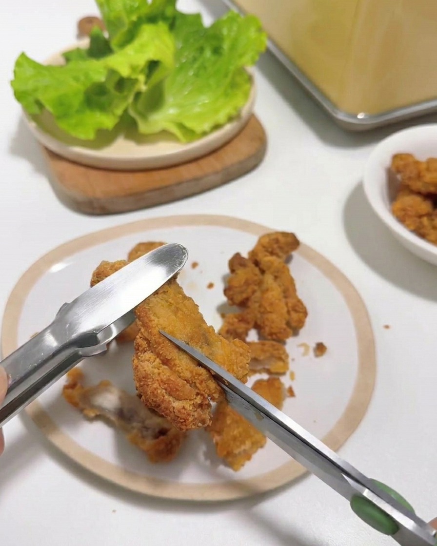 A close-up of kitchen scissors cutting a piece of hot fried chicken held by tongs on a white plate.