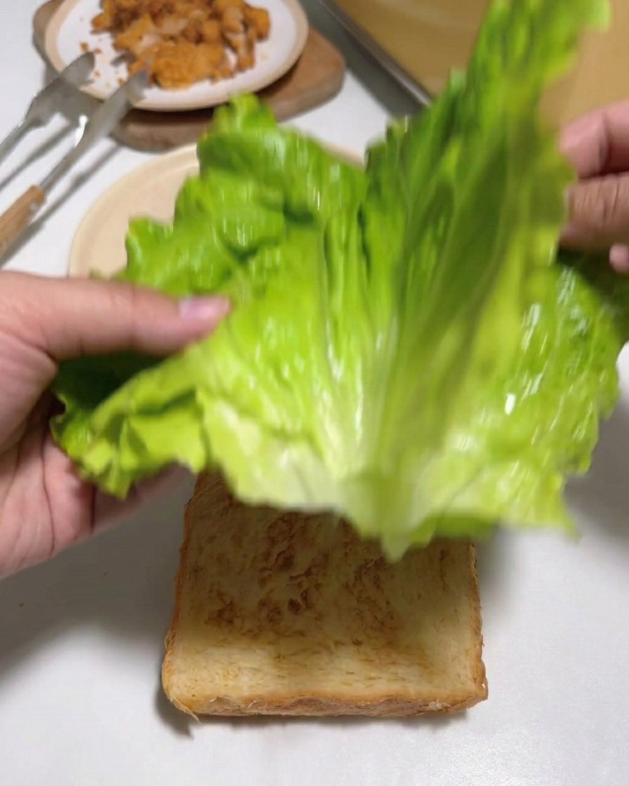 Hands placing large, fresh green lettuce leaves onto a slice of toasted white bread.