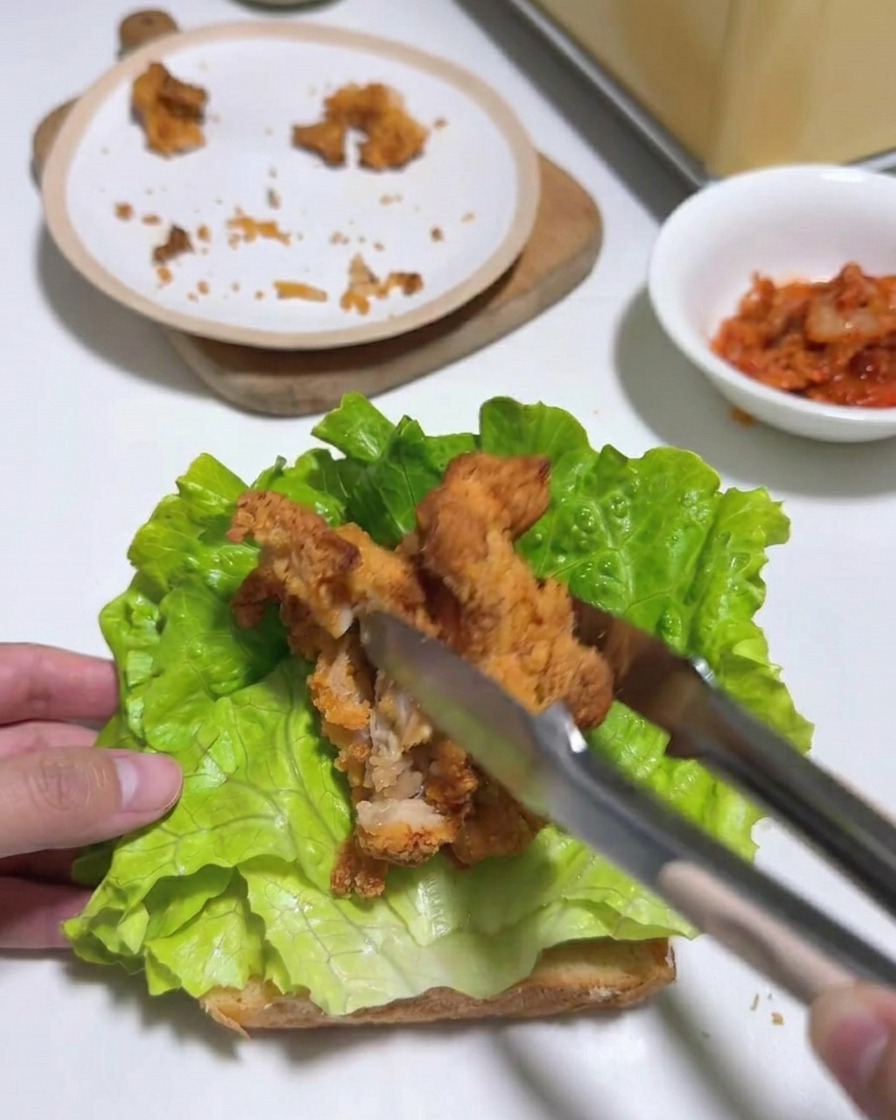 Tongs placing several strips of fried chicken onto a layer of lettuce on toasted bread.