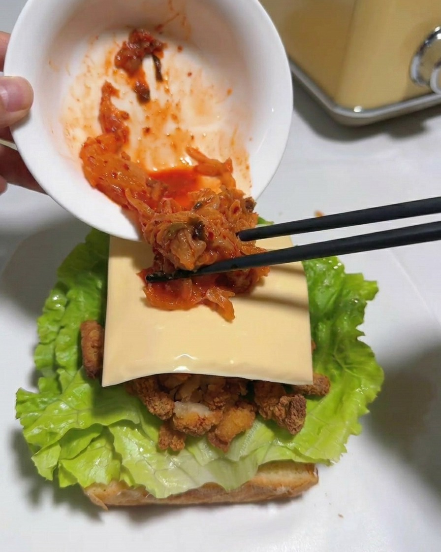 Chopsticks transferring bright red chopped kimchi from a white bowl onto a square slice of cheese over a sandwich.