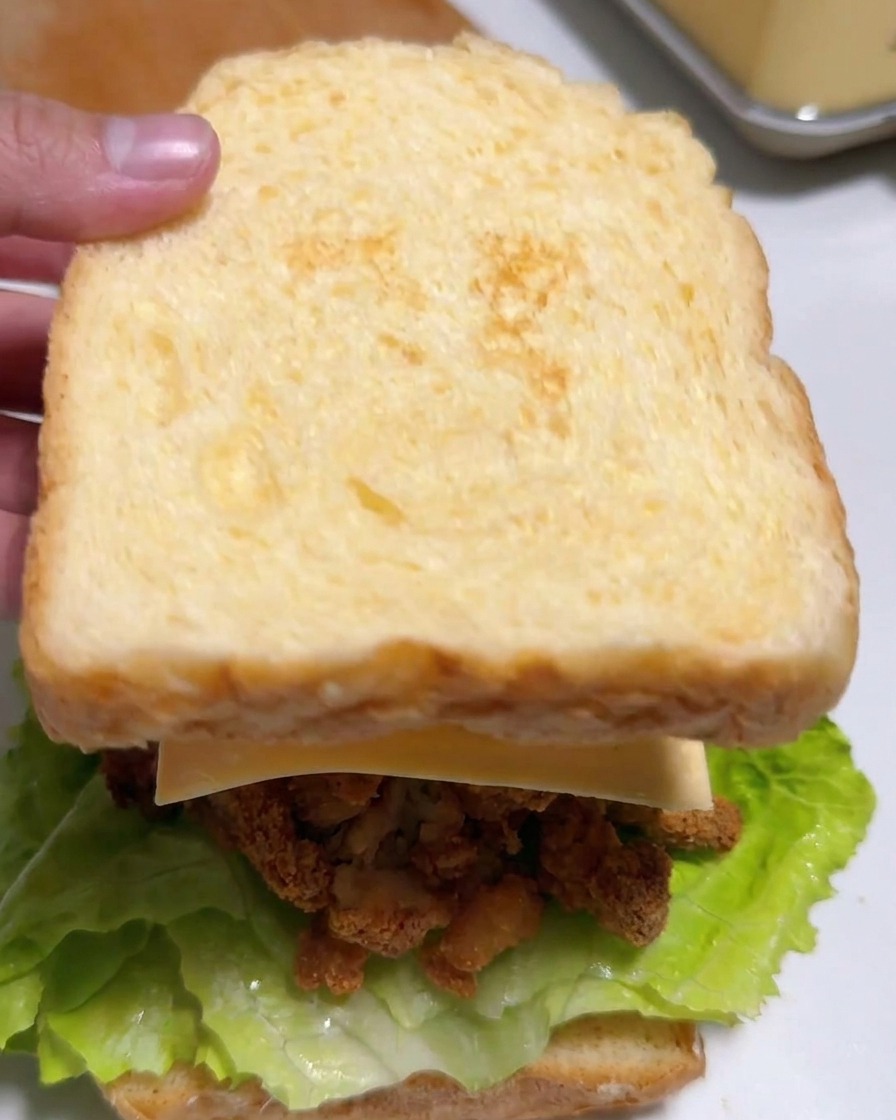 A hand placing a slice of lightly toasted bread on top of a sandwich filled with lettuce, fried chicken, cheese, and kimchi.