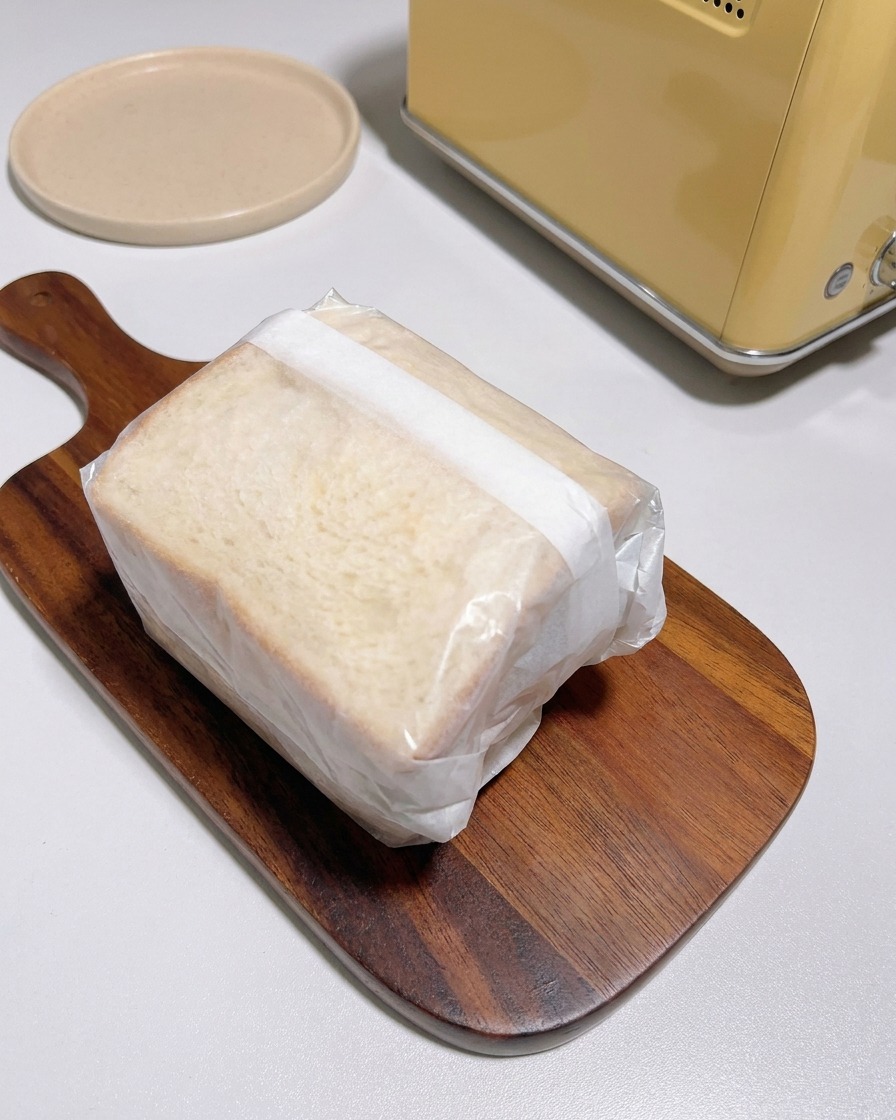 A thick sandwich tightly wrapped in translucent white parchment paper sitting on a wooden serving board next to a yellow toaster.