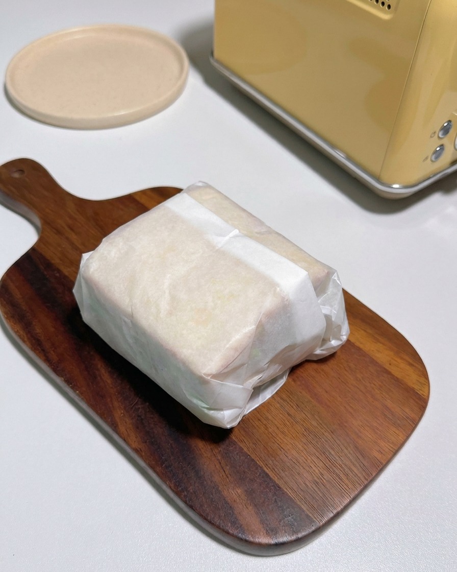Two halves of a parchment wrapped sandwich exposing layers of toasted bread fried chicken cheese and kimchi on a wooden board.