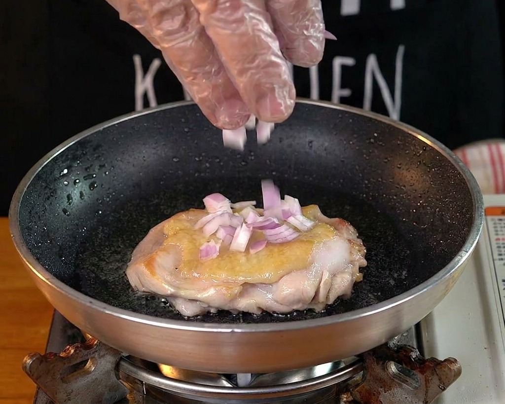 A hand wearing a clear glove sprinkling diced red onions into a pan with a chicken thigh.
