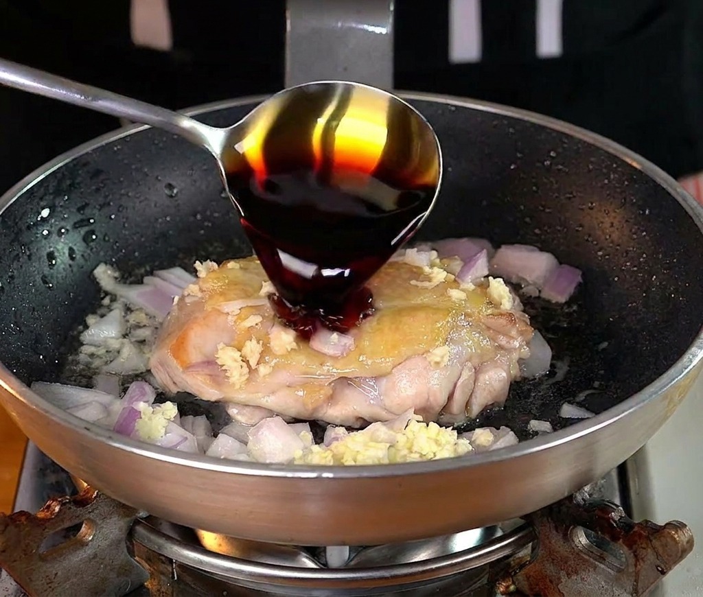 A metal spoon pouring dark soy sauce over a partially cooked chicken thigh in a frying pan.