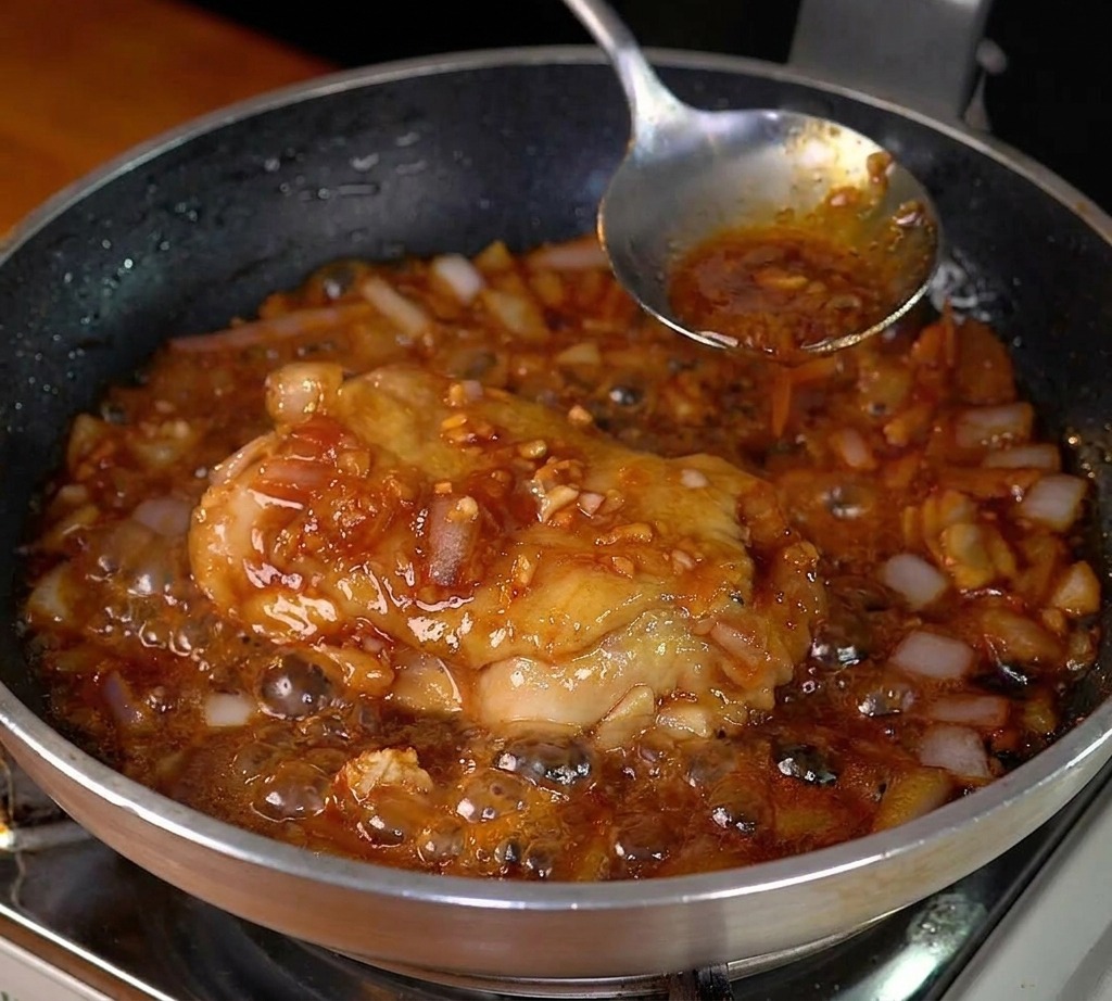 Chicken thigh simmering in a dark, bubbling sauce with softened onions in a stainless steel pan.