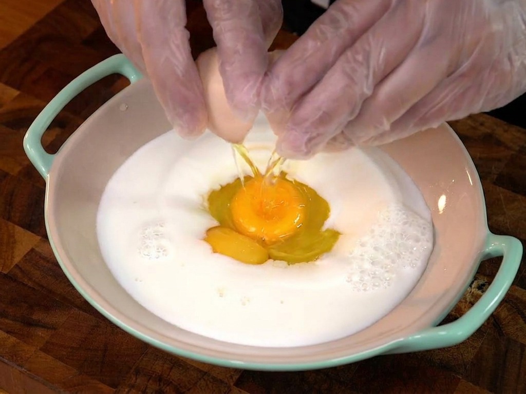 A person cracking an egg into a bowl of milk, with the yolk falling into the center.