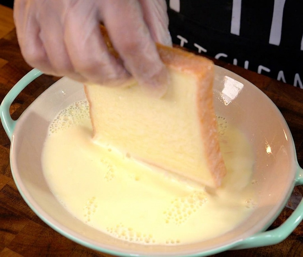 A gloved hand dipping a thick slice of white bread into a shallow bowl filled with a pale yellow egg and milk mixture.