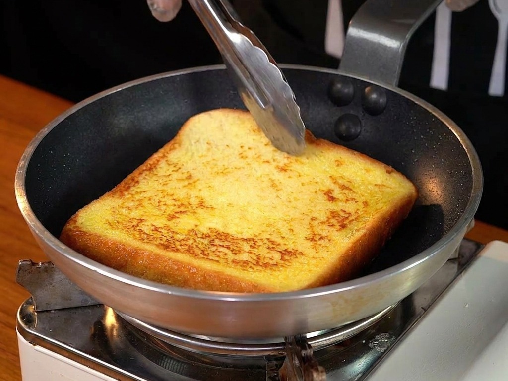 A thick slice of golden-brown French toast being flipped in a frying pan with metal tongs.