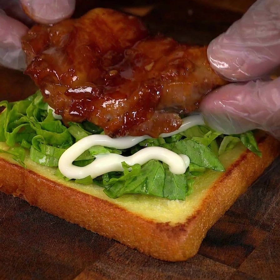 Gloved hands placing a shiny, caramelized chicken thigh onto a sandwich base with lettuce and mayonnaise.