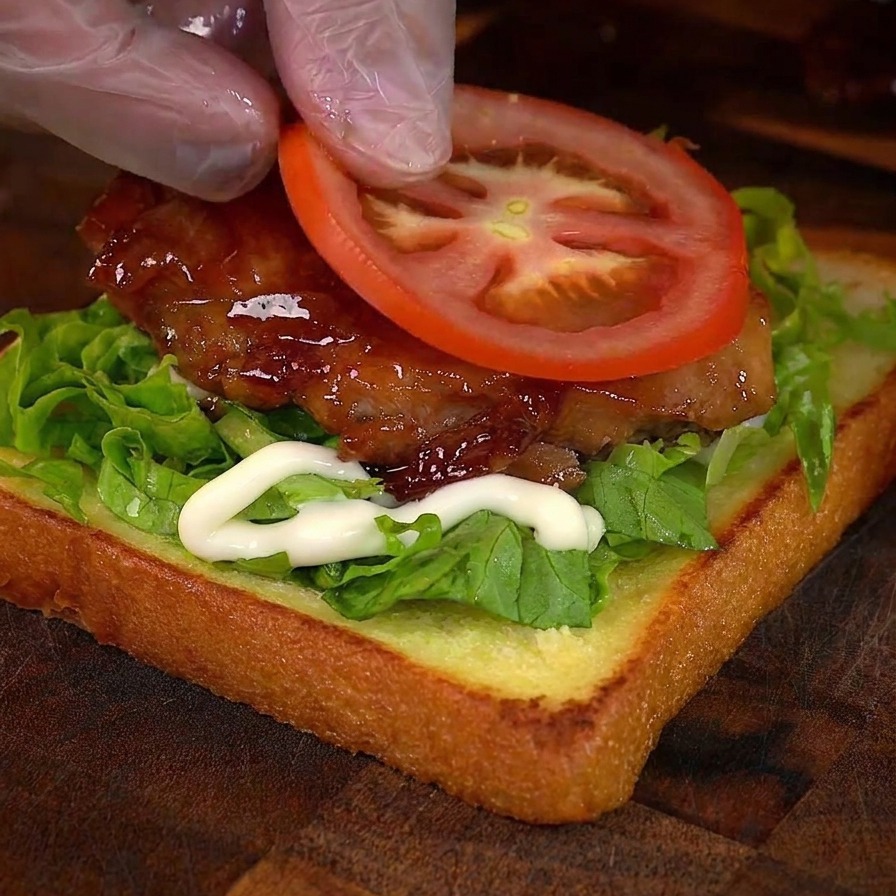 A hand placing a round slice of fresh tomato onto a honey-glazed chicken sandwich.